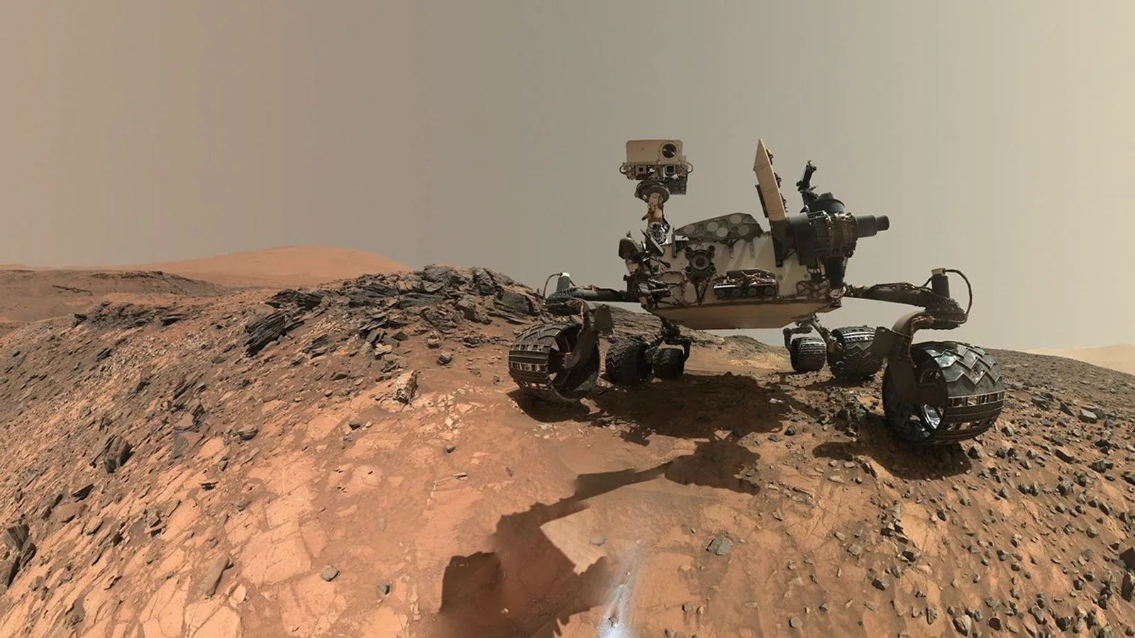 Un rover sobre la superficie marciana, anaranjada y con relieve, bajo un cielo gris plano y rodeado de bloques de roca.