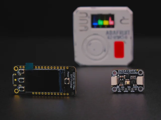 Placa ESP32 S3 con pantalla TFT junto al sensor STCC4 mas SHT41 y el combadge terminado de fondo
