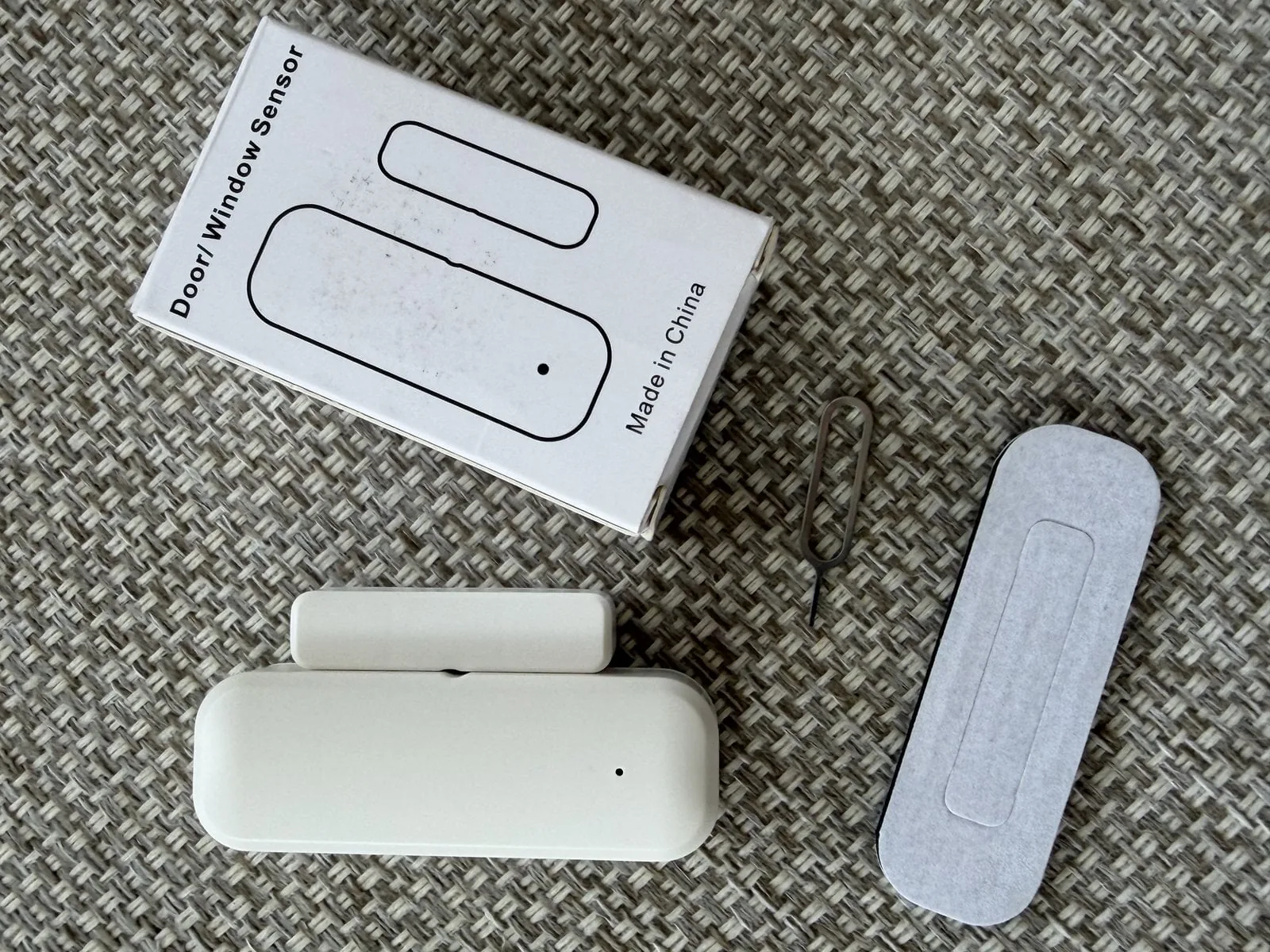 Contacto de puerta/ventana Zigbee con su imán, el empaque y el adhesivo sobre una mesa, sin marca visible