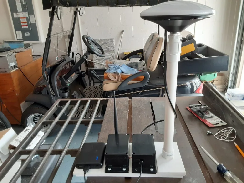 Estación base RTK terminada con antena de grado topográfico sobre mástil, power bank y radio SiK montados en una base de madera